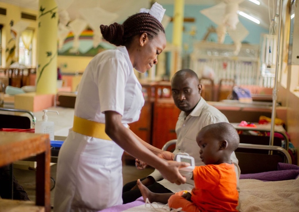 A nurse in a white uniform holds a small monitor to the arm of a child sitting up on a bed in a hospital ward, while a man sitting beside the bed watches. Painted murals and further beds fill the room behind them.