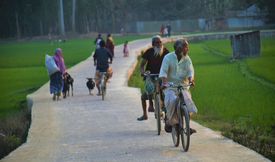 People walking and cycling along a narrow concrete path raised between flooded green paddy fields in the late afternoon. Two men ride bicycles towards the camera; behind them a woman carries a sack on her head and two goats follow.